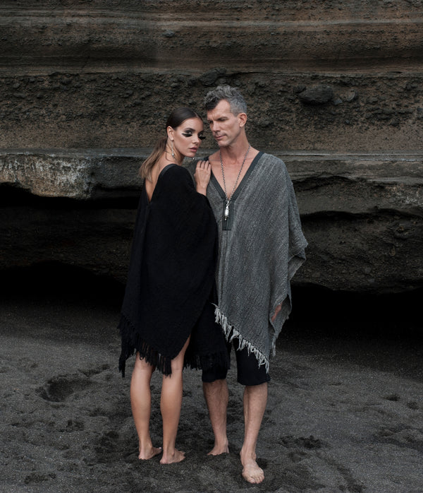 Two people standing on a beach wearing a black poncho and the other person wearing a grey poncho with rocky cliffs in the background